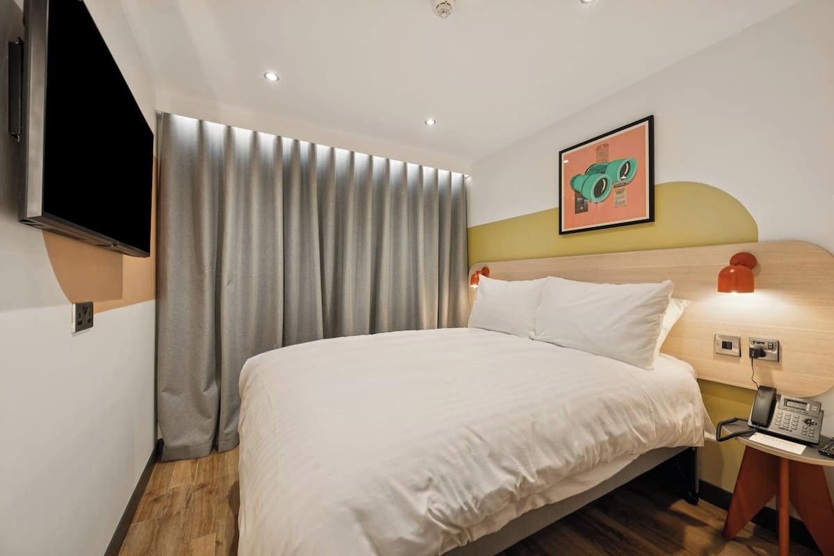 Quarto de hotel com cama, televisão na parede, telefone e pintura na parede.
