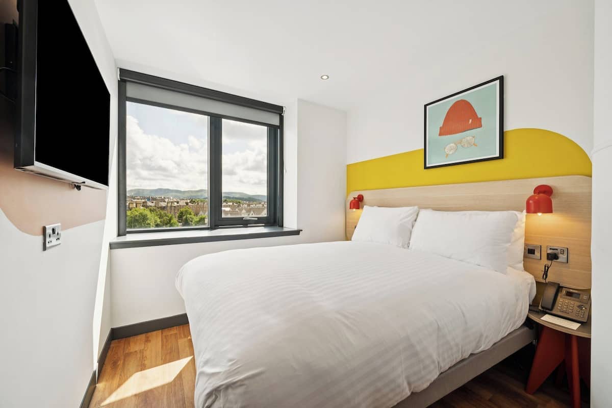 Quarto com cama, janela com vista para a cidade, TV na parede e telefone ao lado da cama.