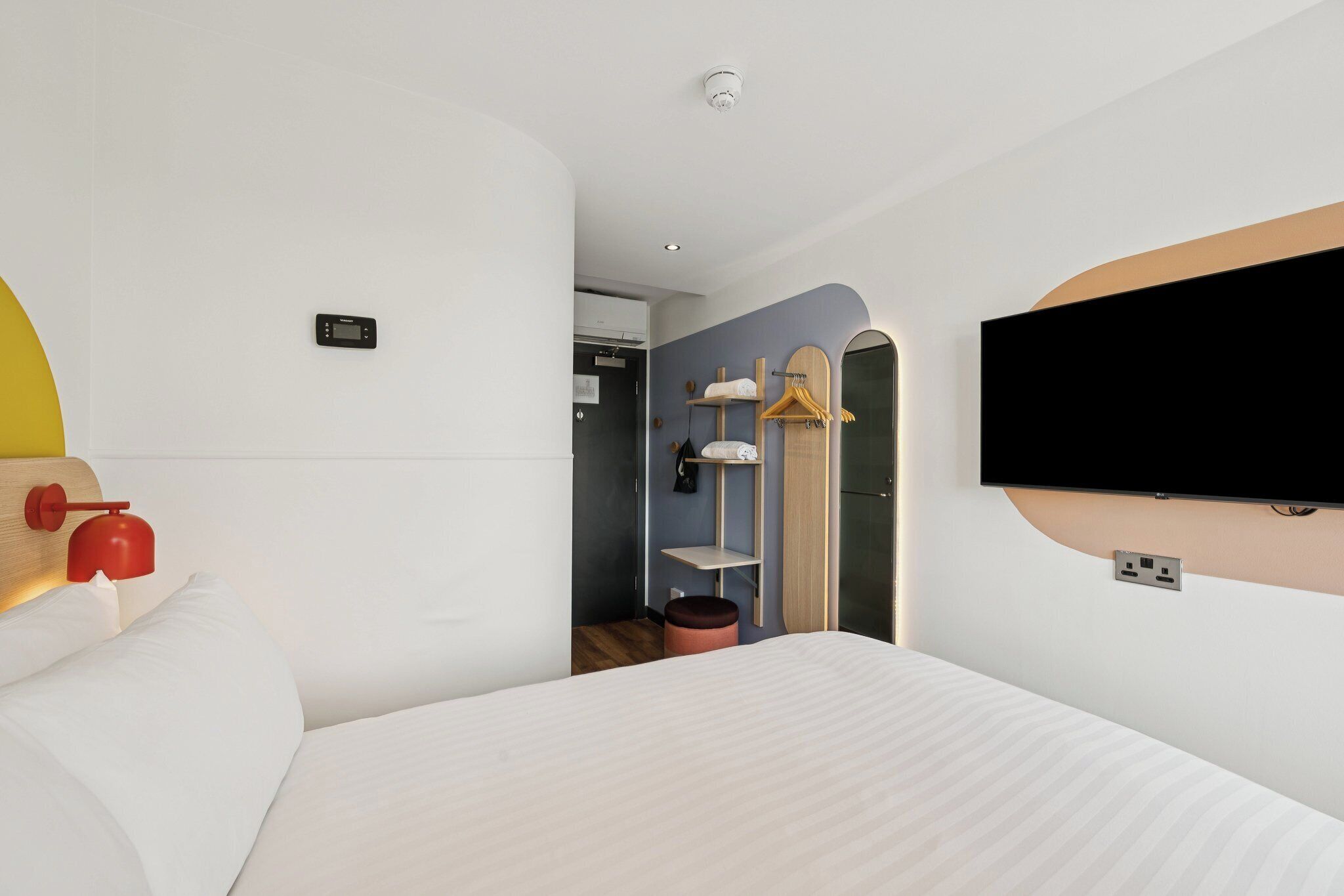 Een moderne hotelkamer met een groot bed, een flatscreen televisie aan de muur, een rode wekker en een kleine digitale thermostaat.
