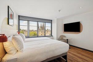 A hotel room with a large window, a bed, a chair, a small table, and a mounted TV.
