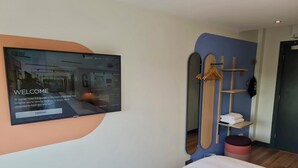 A hotel room with a large screen displaying a welcome message, a bed, a wooden shelf with towels, and a mirror with a wooden frame. - Garner Edinburgh Haymarket by IHG (Edinburgh)