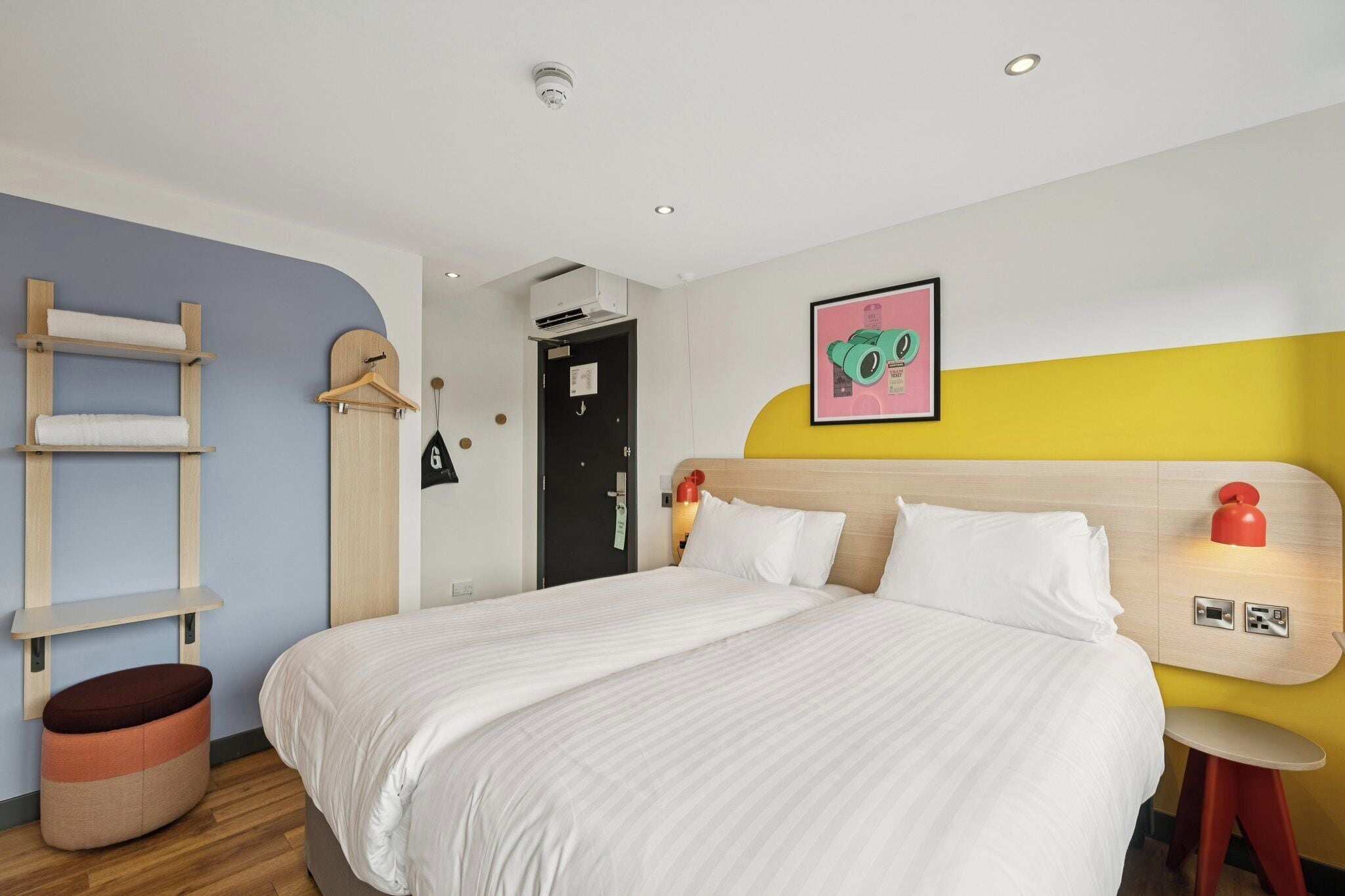 A hotel room with a bed, a wooden ladder, a hanging coat rack, and a wall-mounted light fixture.