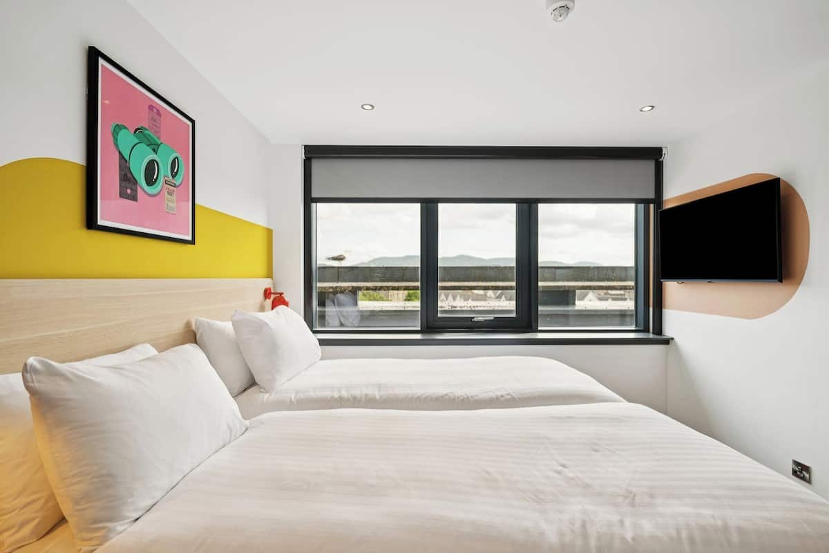 Quarto de hotel moderno com uma cama grande, janela com vista e televisão na parede.