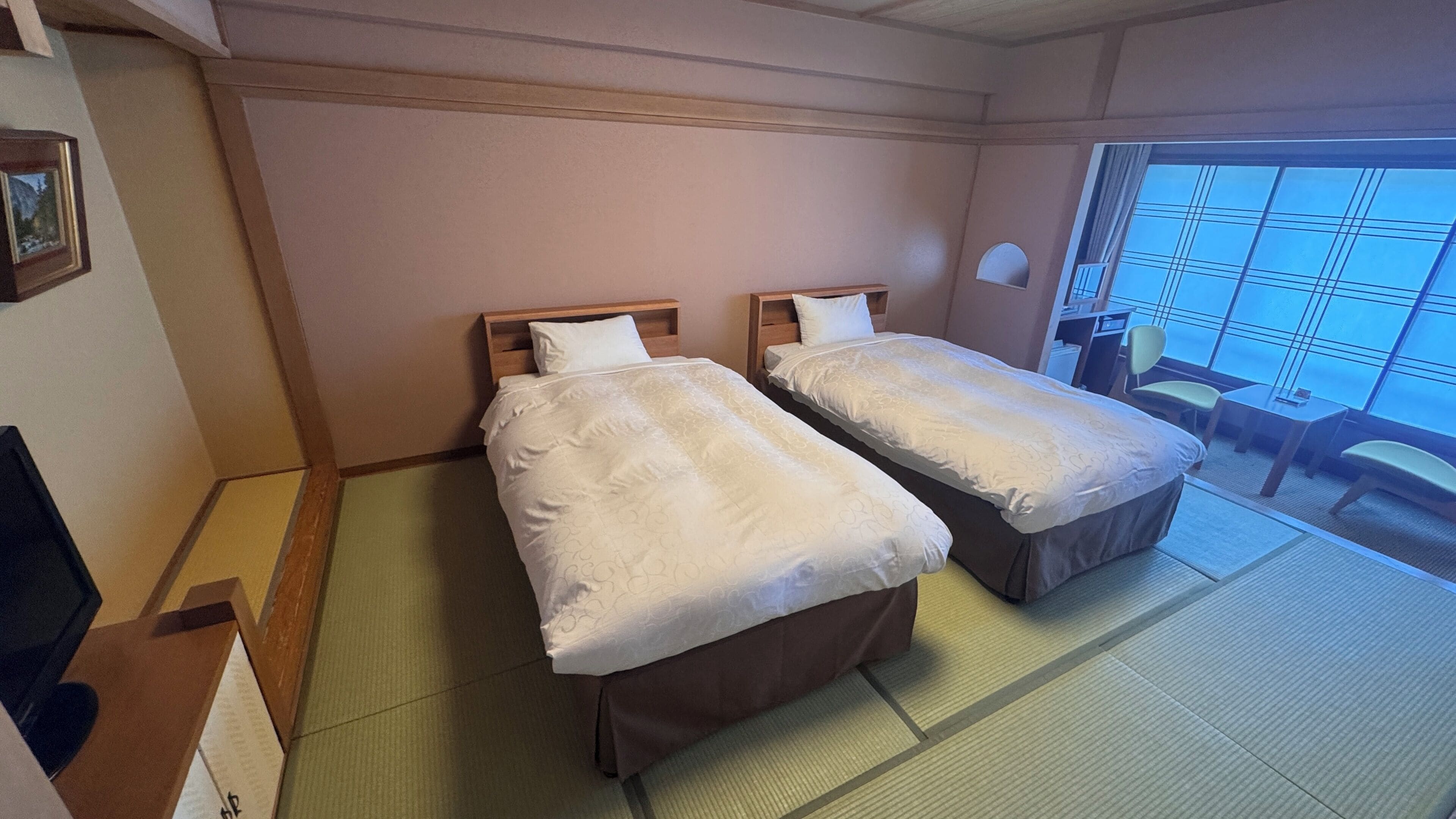 A hotel room with two beds, a small table, a chair, and a window with a view.
