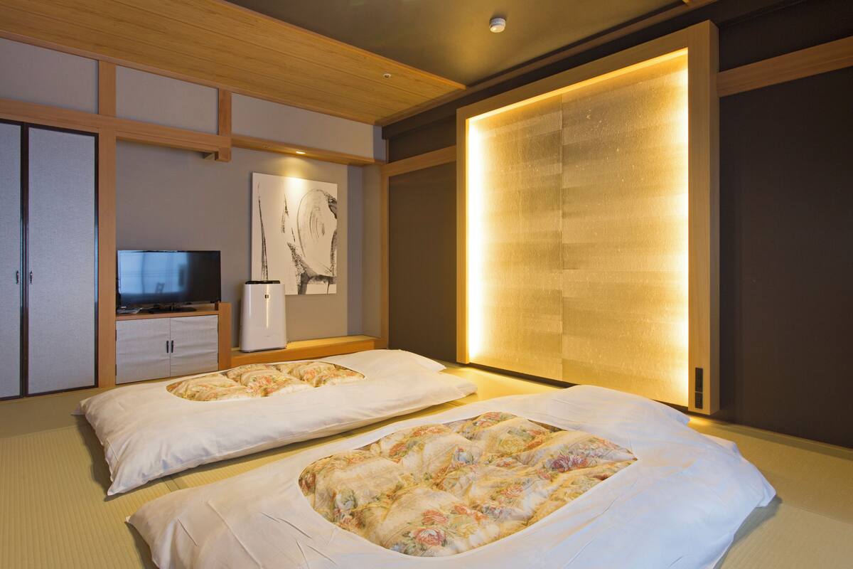 A hotel room with two beds, a television, artwork on the wall, and a sliding door with a warm light.