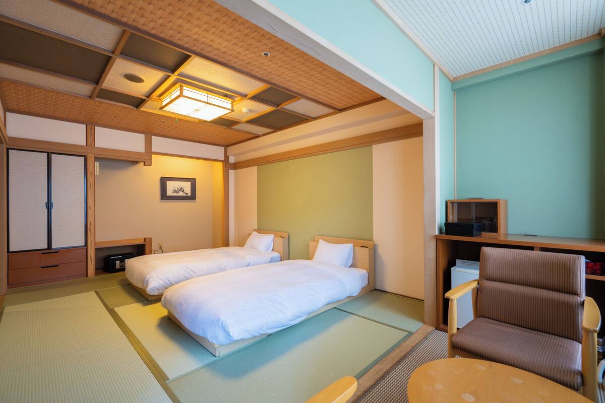 A traditional Japanese-style room with tatami flooring, two beds, a wooden chair, and a shelf with a television.
