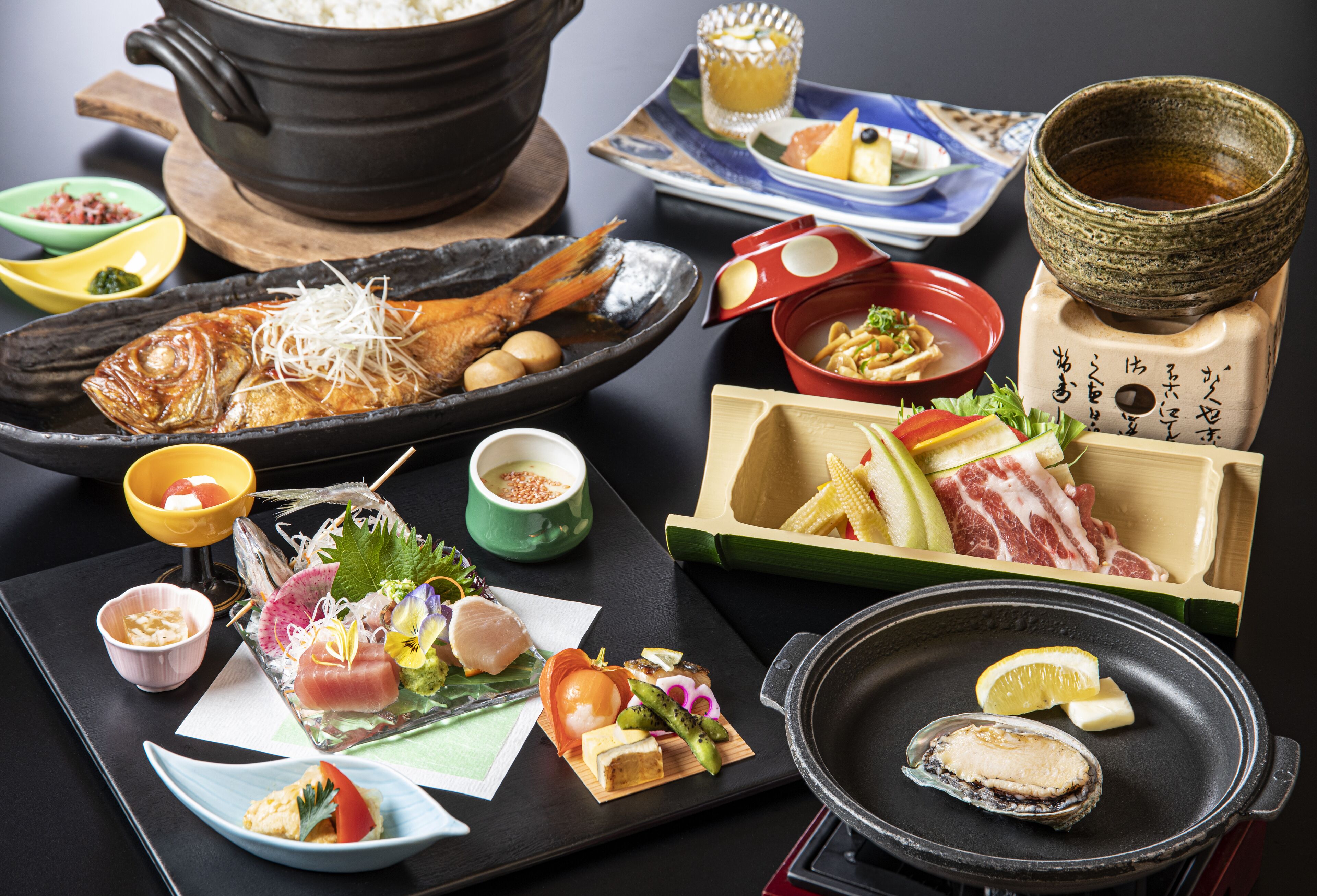 Tarjolla on monenlaisia japanilaisia ruokia, kuten sushia, grillattua kalaa ja kasviksia, jotka on aseteltu mustalle tarjottimelle.