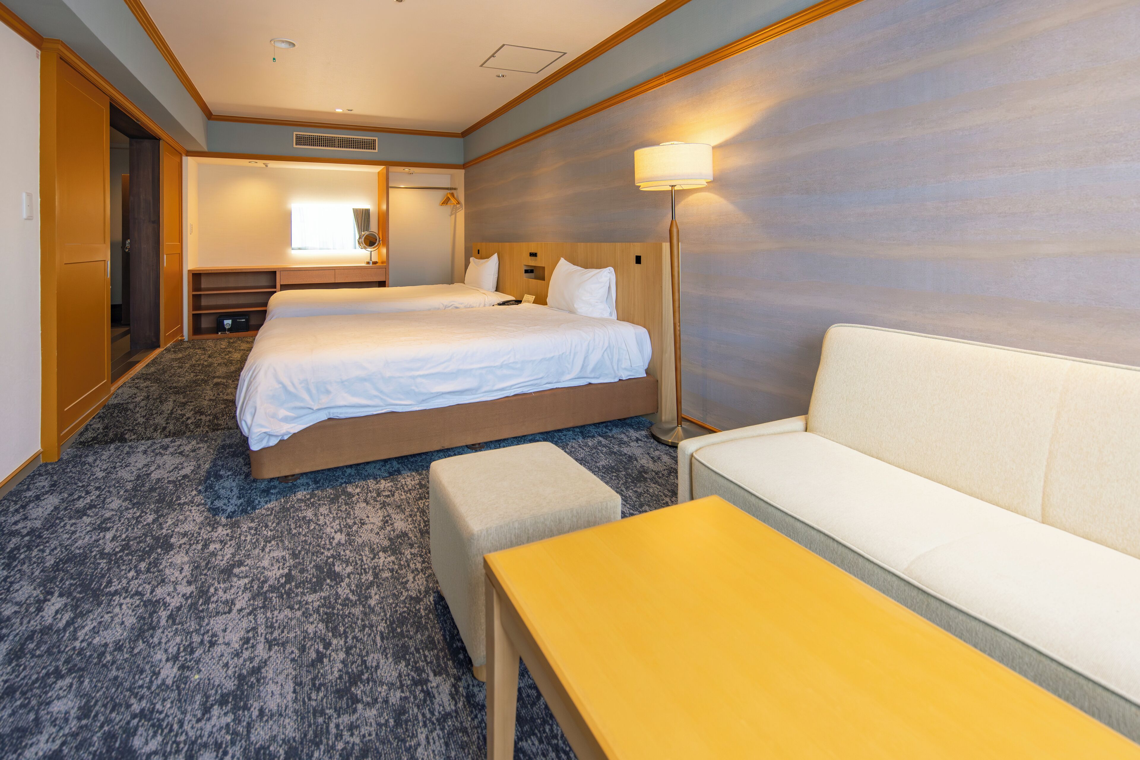 A hotel room with two beds, a sofa, a small table, a mirror, and a lamp.