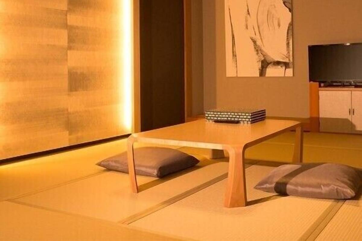 A room with tatami flooring, a low wooden table, cushions, a television, and a painting on the wall.
