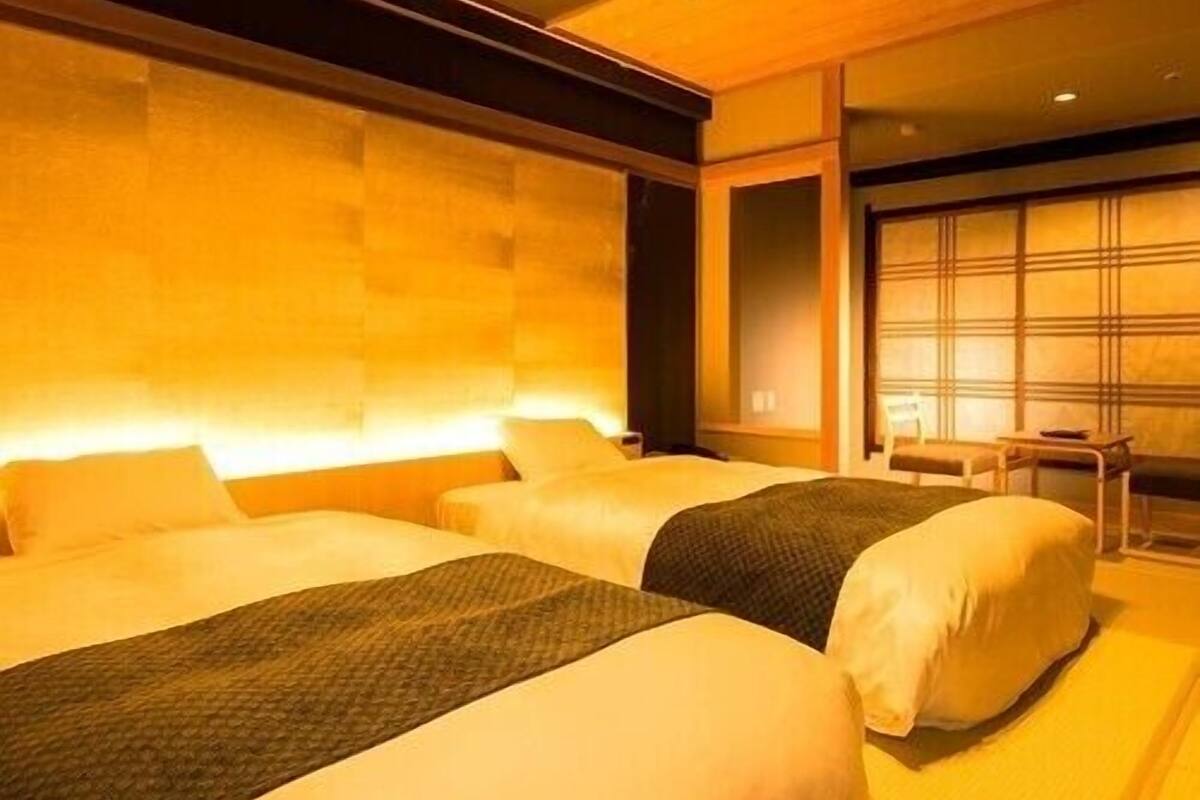 A hotel room with two beds, a wooden headboard, a small table, and a window with blinds.