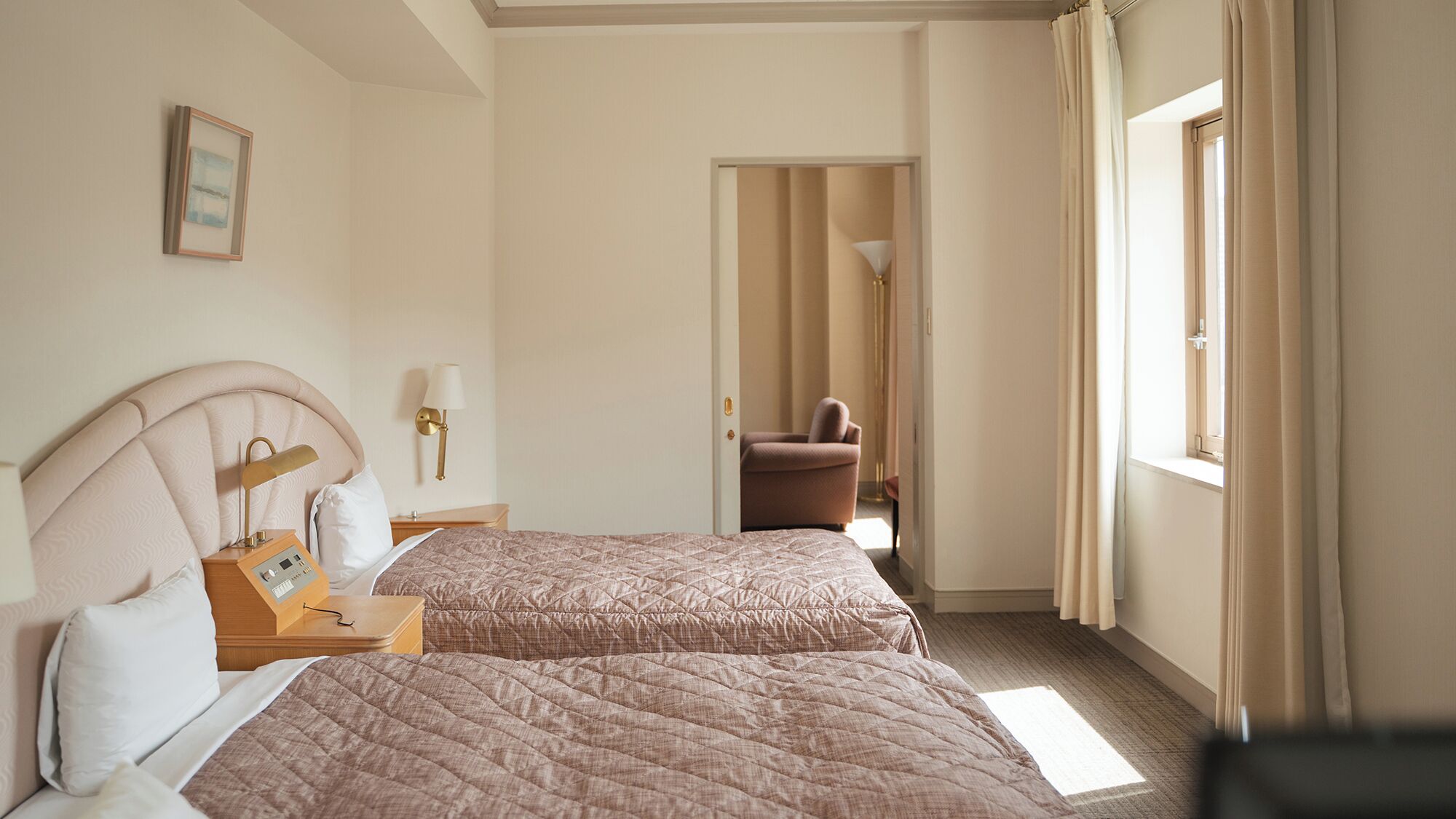 Hotellihuone, jossa on kaksi sänkyä, yöpöytä lampun kanssa, seinällä oleva kehystetty kuva ja näkymä käytävälle.
