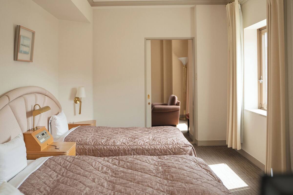 A hotel room with two beds, a bedside table with a lamp, a framed picture on the wall, and a view of the hallway.