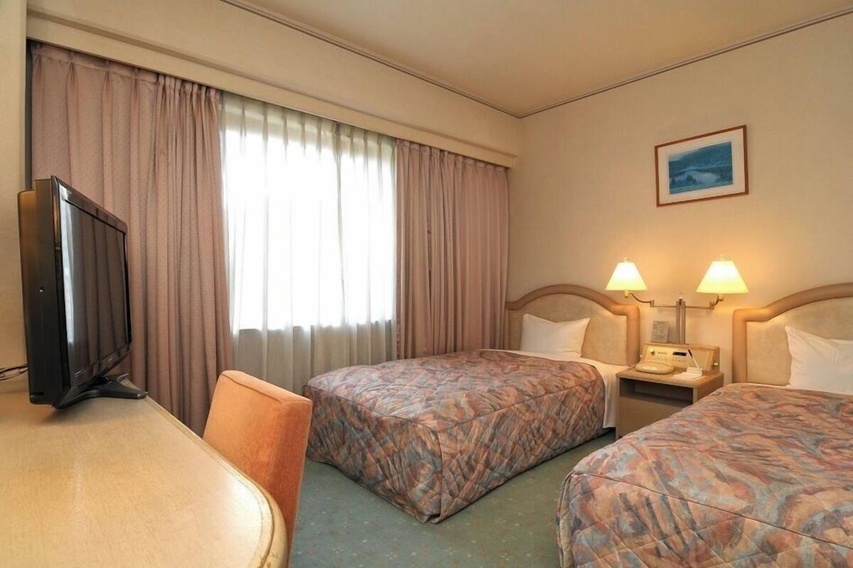 A hotel room with two beds, a TV, a desk, and a window with curtains.