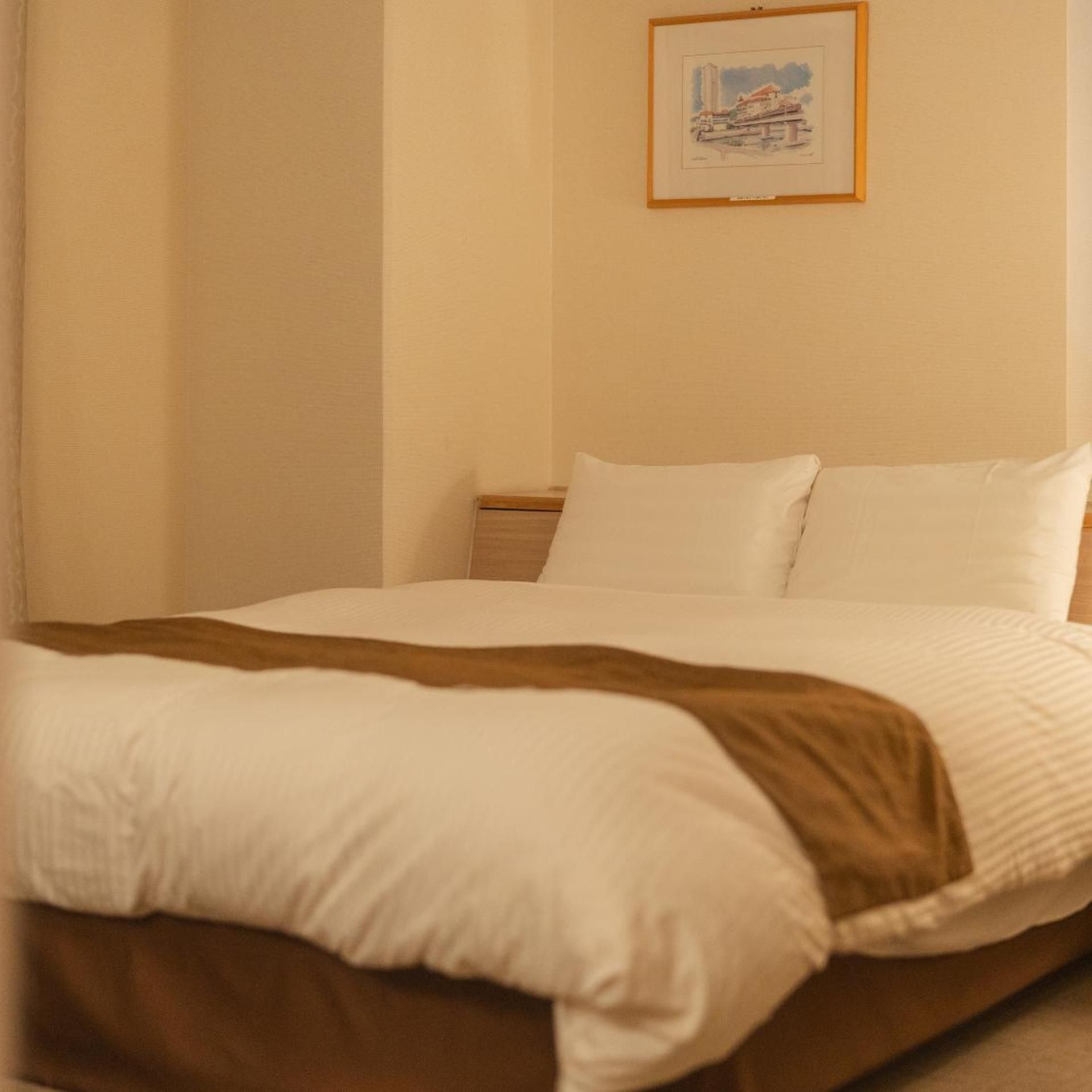 Een enkel bed met witte beddengoed en een bruine plaid, een ingelijst foto aan de muur en een houten nachtkastje.