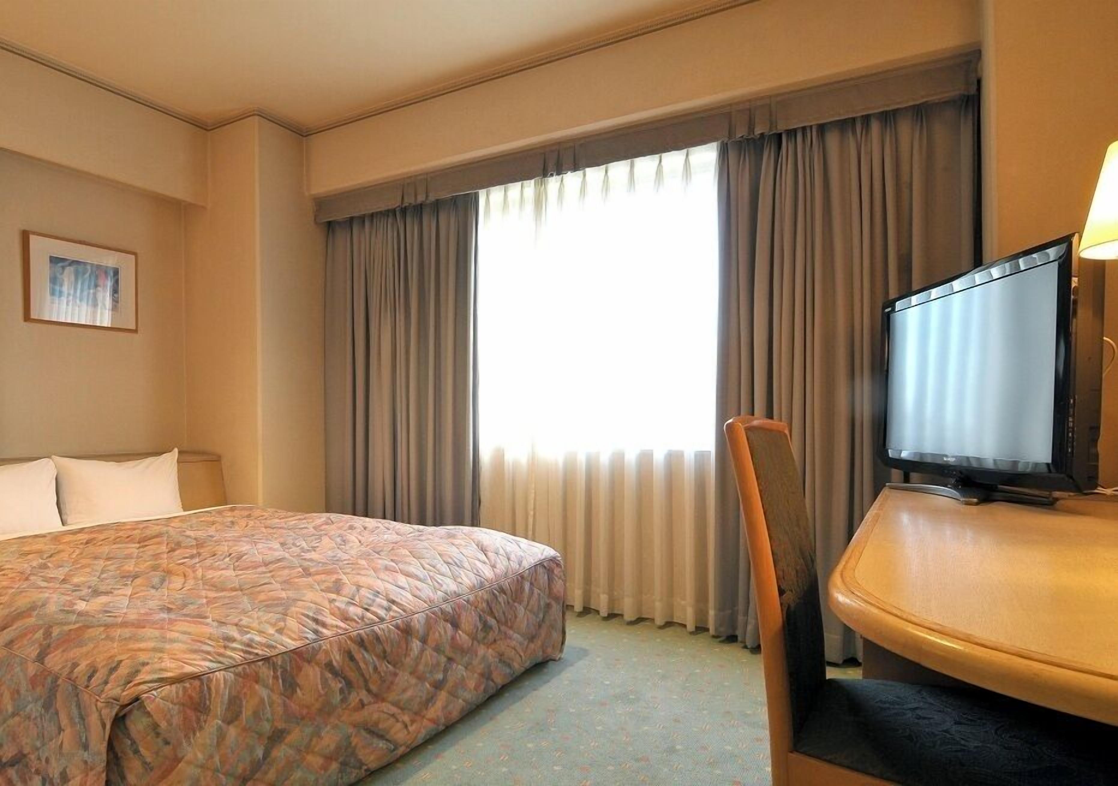 Una habitación de hotel con cama, televisión, silla y ventana con cortinas.