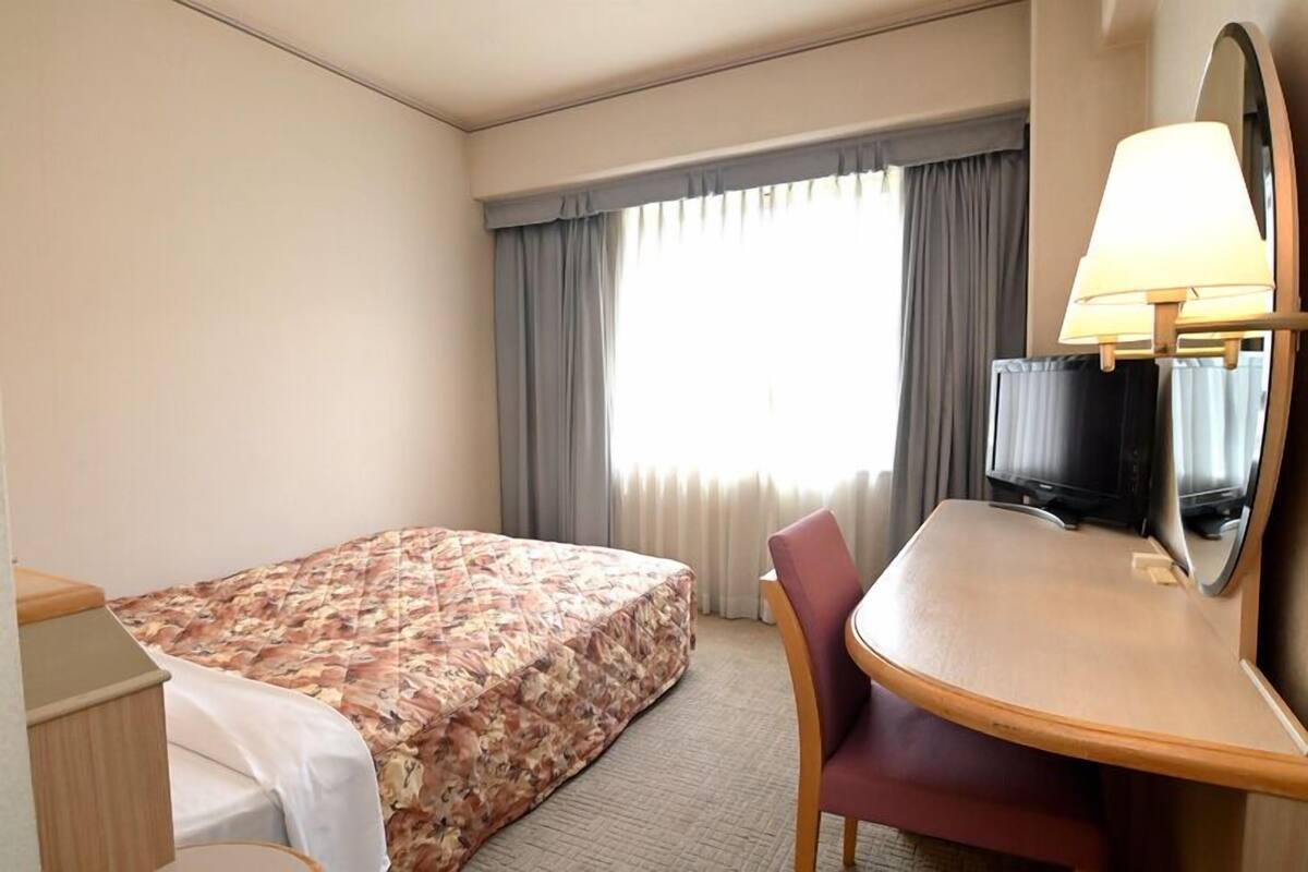 A hotel room with a bed, a desk with a chair, a television, and a window with curtains.
