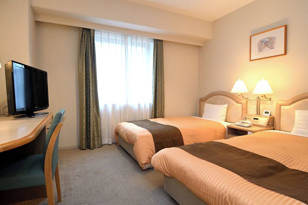Una habitación de hotel con escritorio, dos camas, televisor y ventana con cortinas.