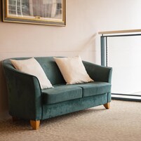Un sofá color turquesa con patas de madera y dos cojines blancos en una habitación con alfombra estampada.