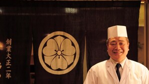 A person in a white chef's uniform standing in front of a room with a dark curtain featuring a large circular design and Japanese text.