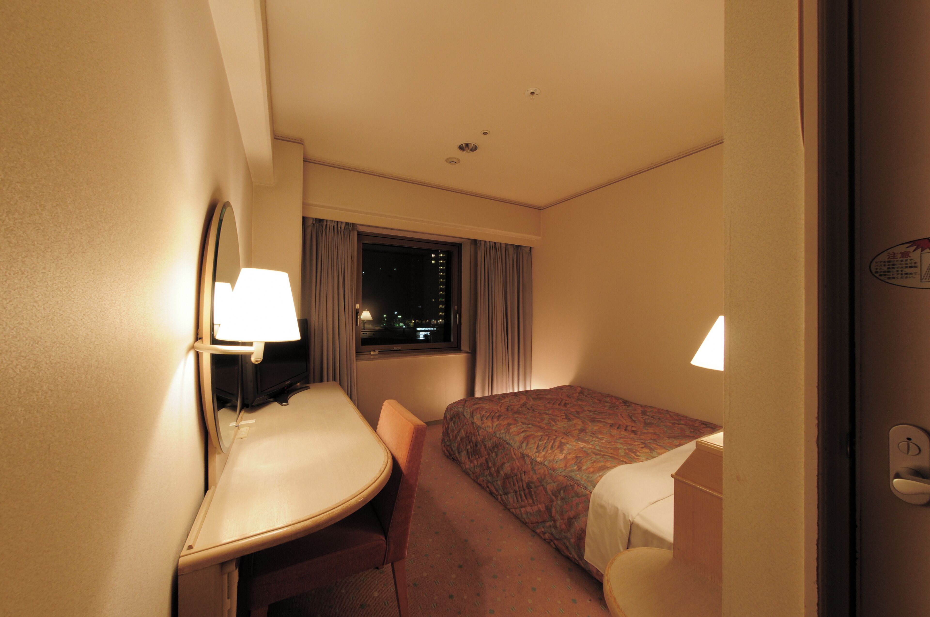 Habitación de hotel con cama, escritorio, silla y ventana con cortinas, mostrando una vista nocturna al exterior.
