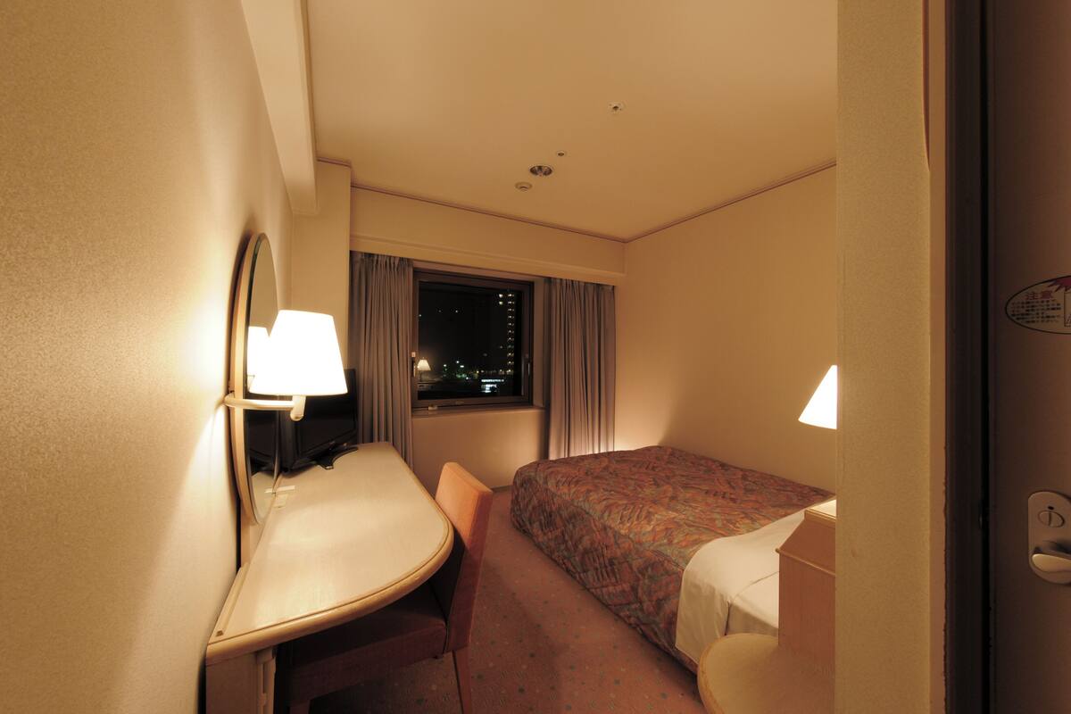 A hotel room with a bed, a desk, a chair, a window with curtains, and a night view outside.