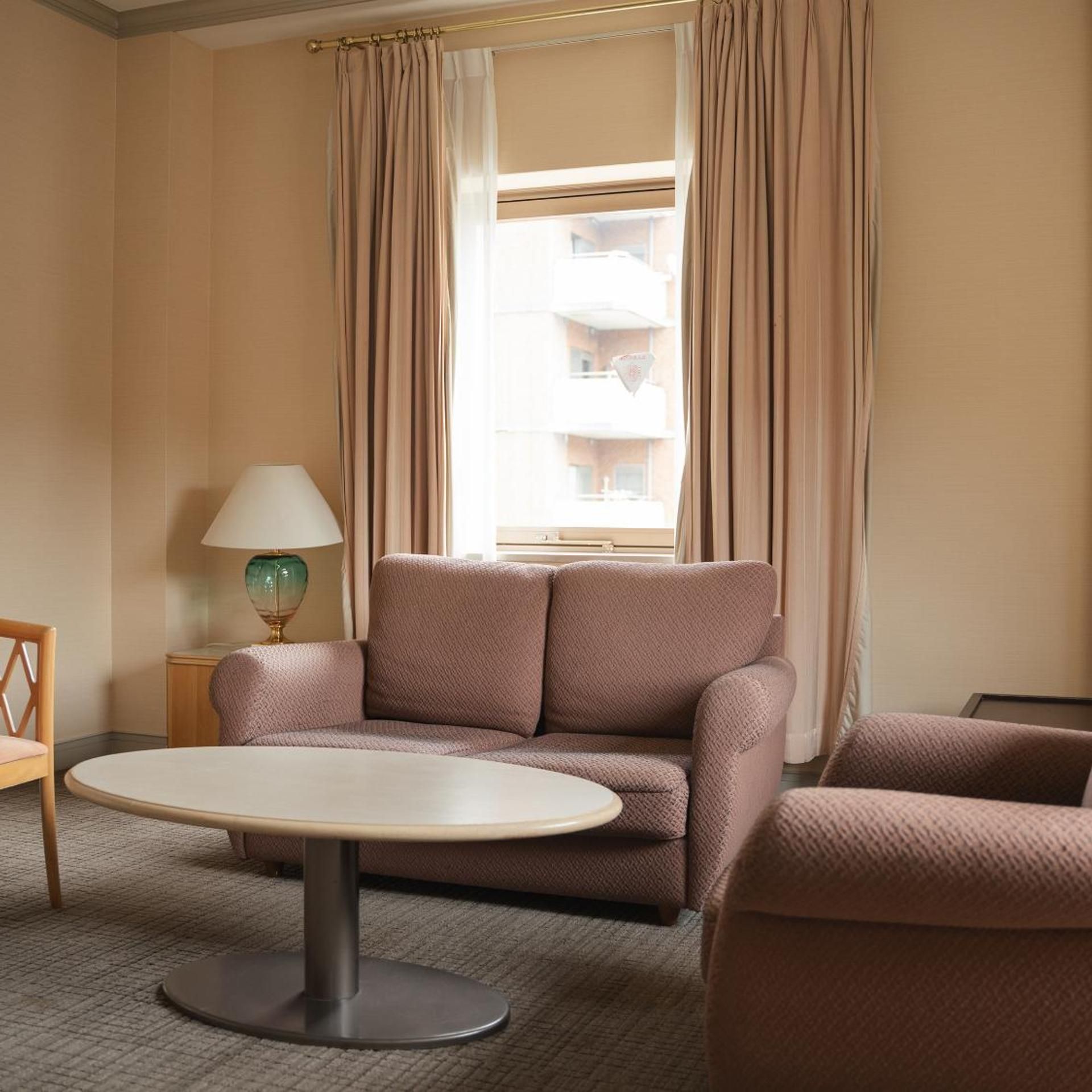 A hotel room with a sofa, a round table, a lamp, and a view of a building through the window.