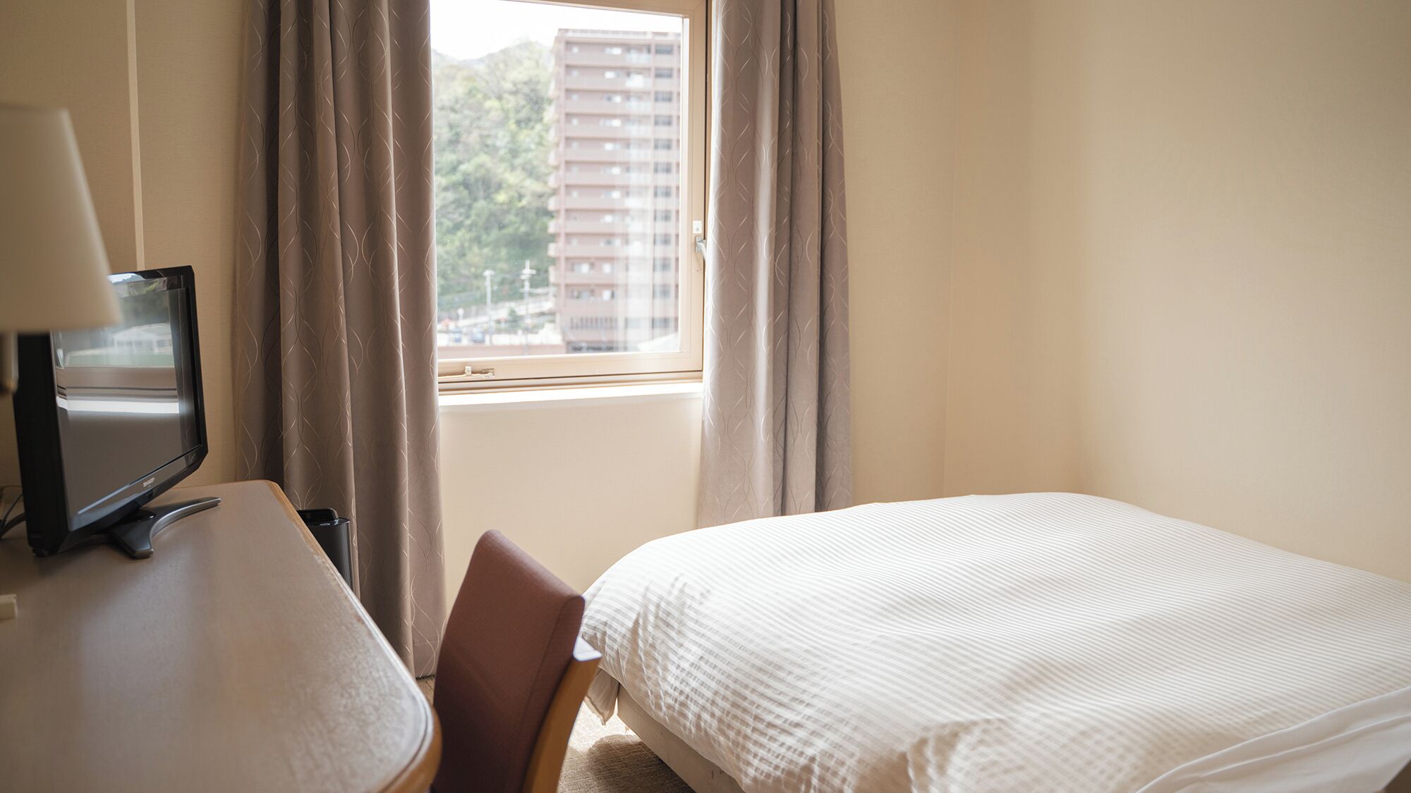 Hotellihuone, jossa on sänky, työpöytä lampun kanssa, tuoli ja ikkuna, jossa on verhot.
