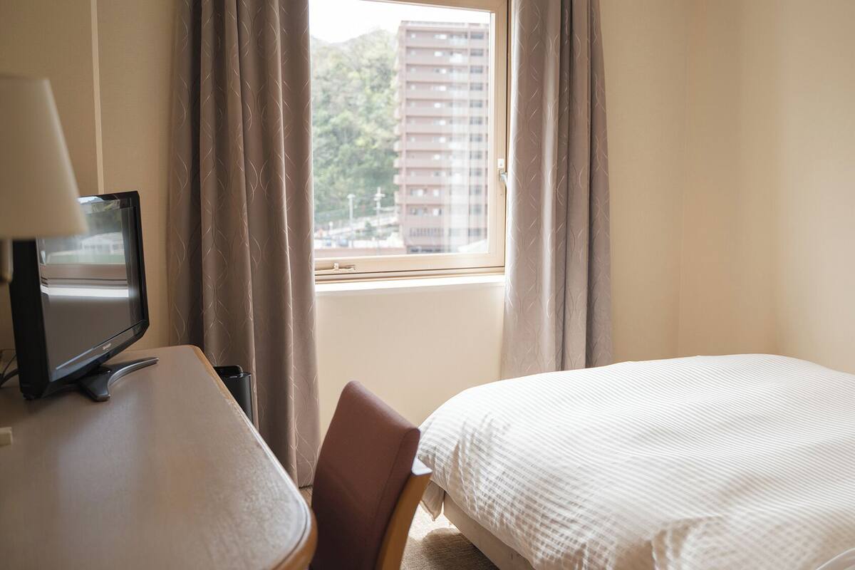 A hotel room with a bed, a desk with a lamp, a chair, and a window with curtains.