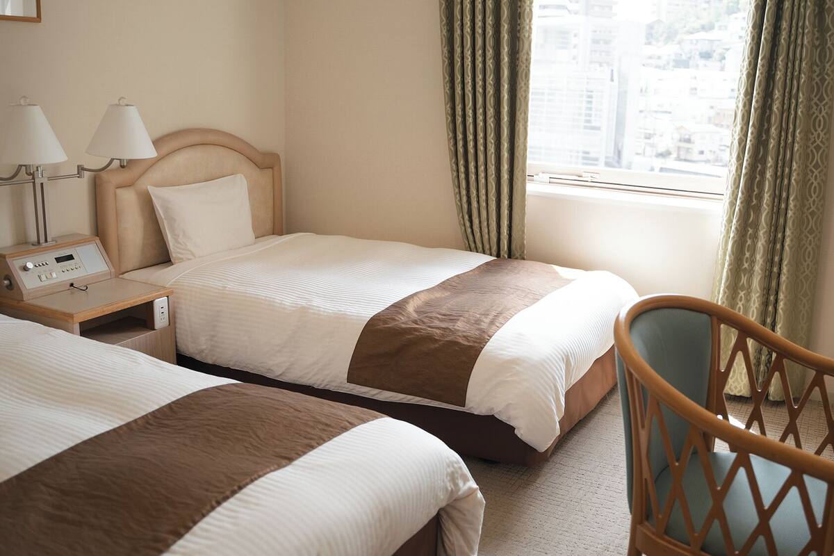 A hotel room with two beds, a bedside table with lamps, a chair, and a view of the city through the window.