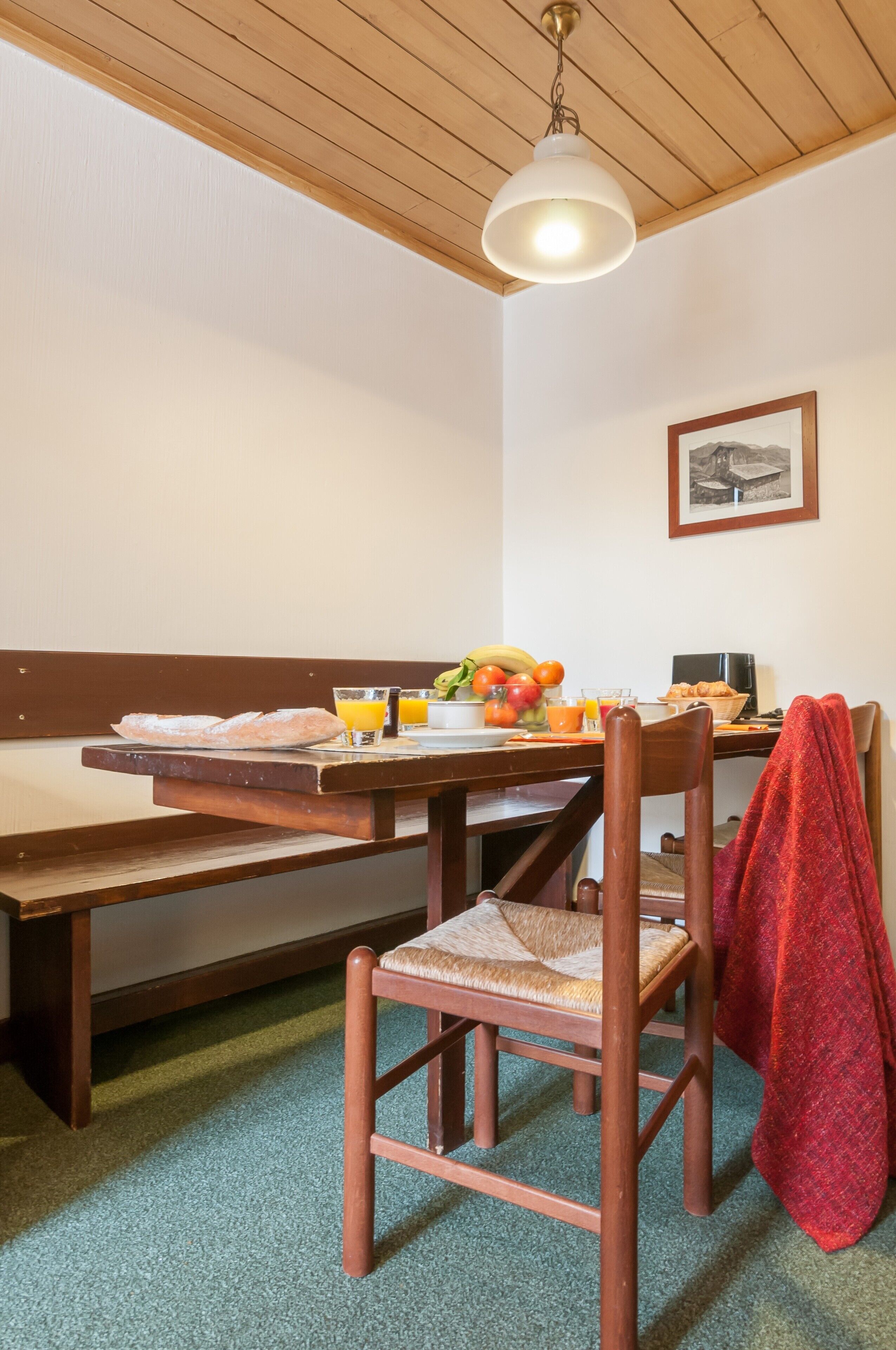 A dining area with a wooden table, bench, and chairs. There is a fruit bowl with bananas, oranges, and apples, and a glass of orange juice. A red towel is draped over one chair.