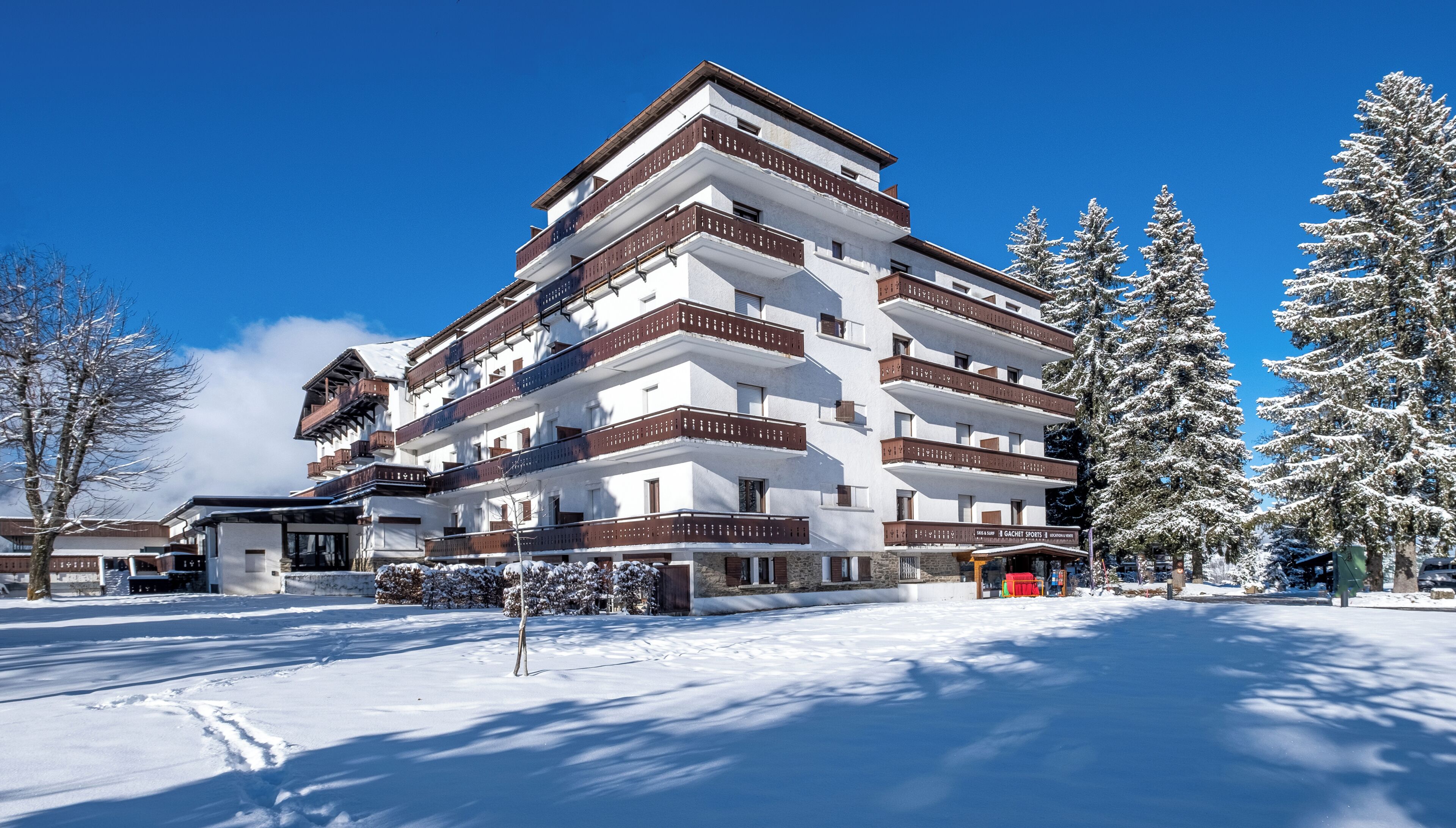 A multi-story hotel with balconies, snow-covered trees, and a clear blue sky.