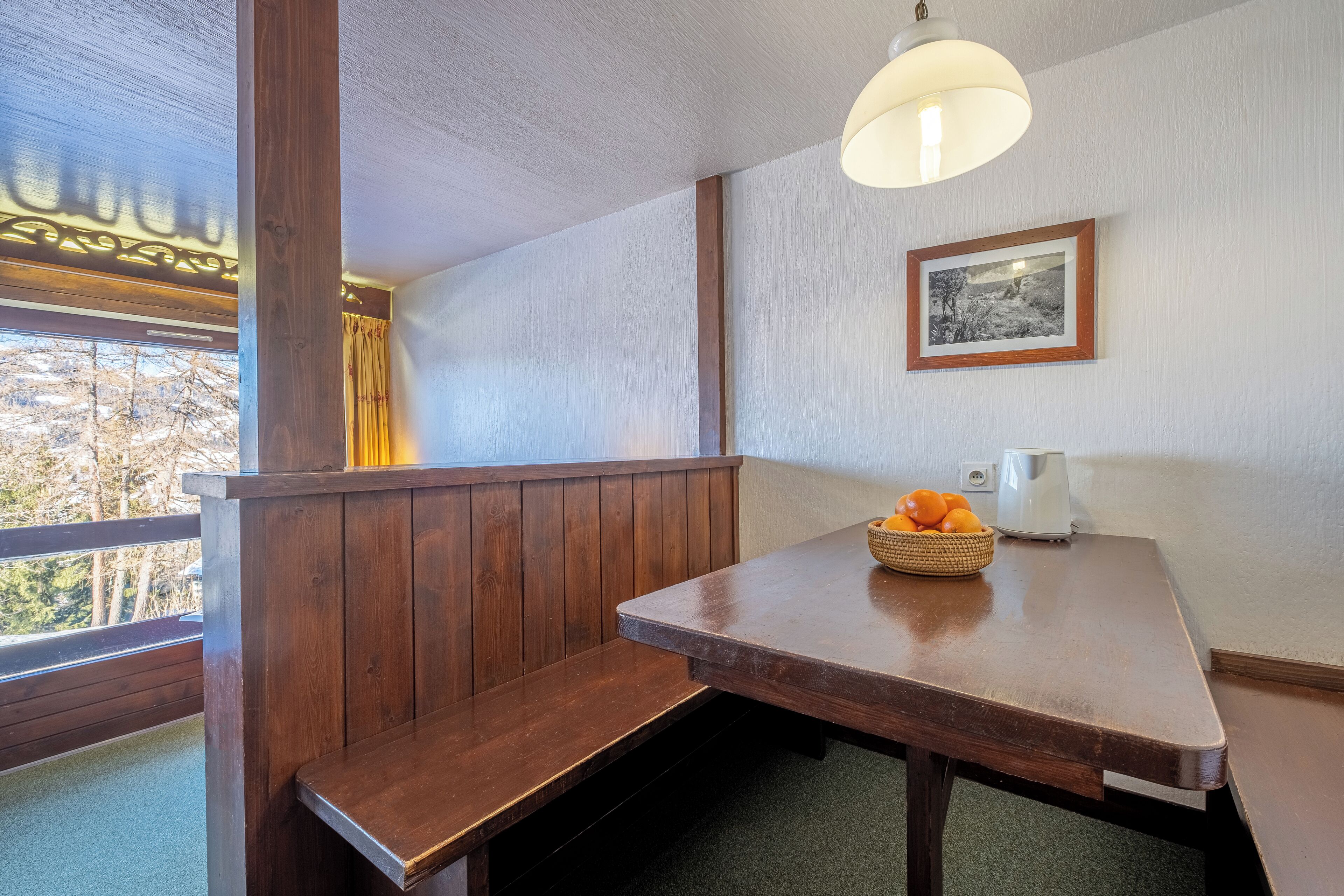 A wooden bench and table in a room with a window, a framed picture on the wall, and a basket of oranges on the table.