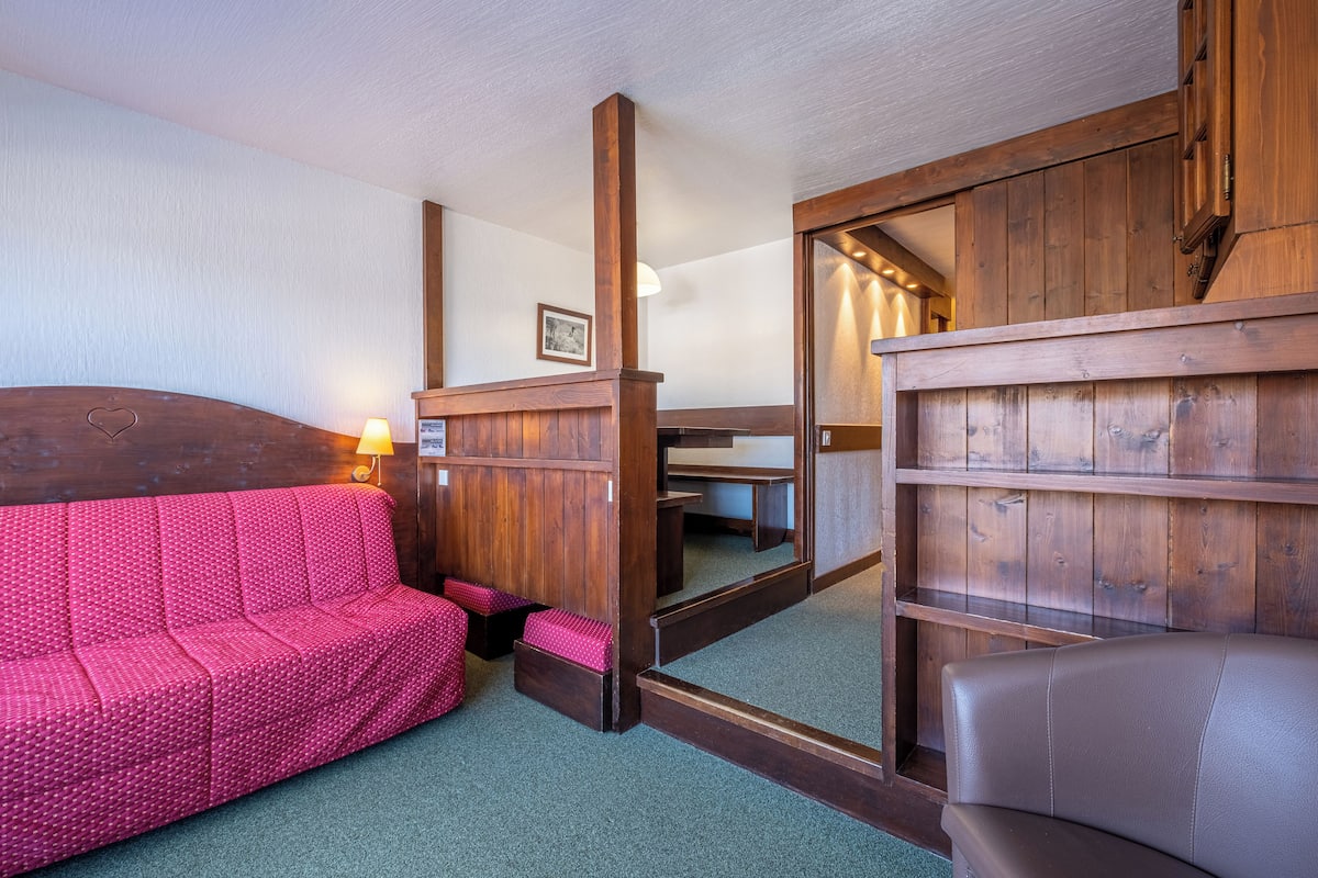 Ein gemütliches Zimmer mit einem Holzkopfstück, einem rosa gemusterten Sofa, einem braunen Ledersessel und einem Spiegel, der den Raum reflektiert.
