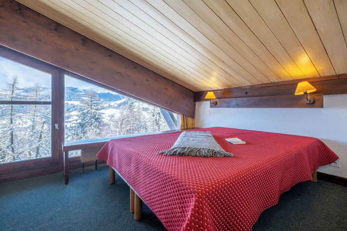 Ein Schlafzimmer mit großem Fenster, Holzboden und einer roten Tagesdecke.