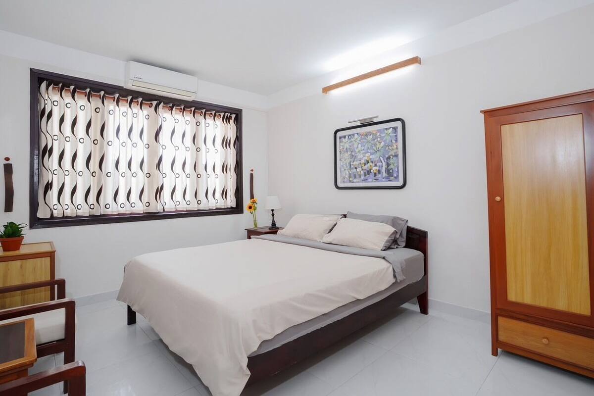 A bedroom with a bed, a nightstand, a potted plant, a window with curtains, a framed picture on the wall, and a wardrobe.