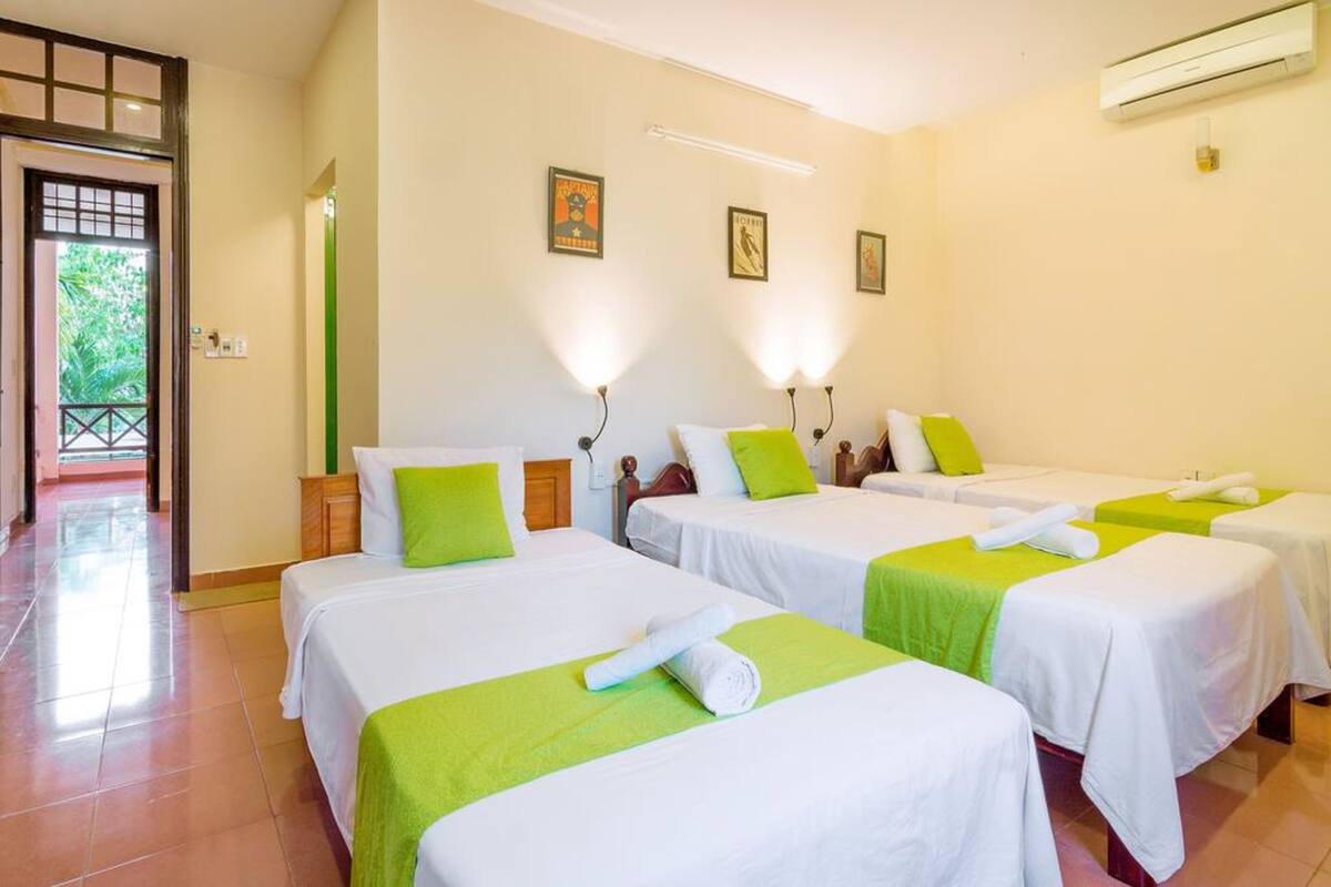 A hotel room with four single beds, each with a green bedspread and white linens. There are wall-mounted lights above each bed, and a door leading to a balcony.