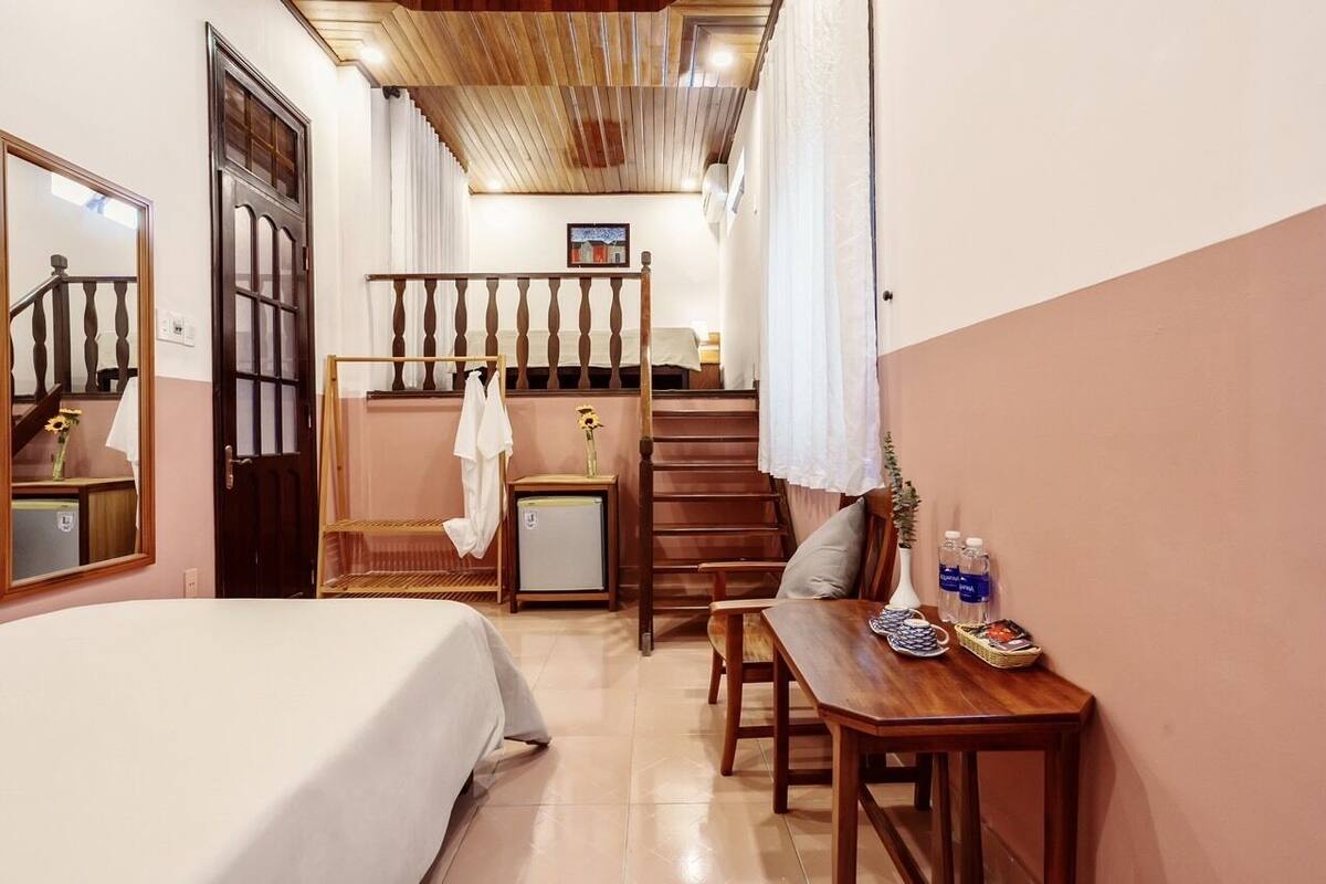 A hotel room with a bed, a wooden table with a vase and bottles, a small fridge, and a staircase with a railing.