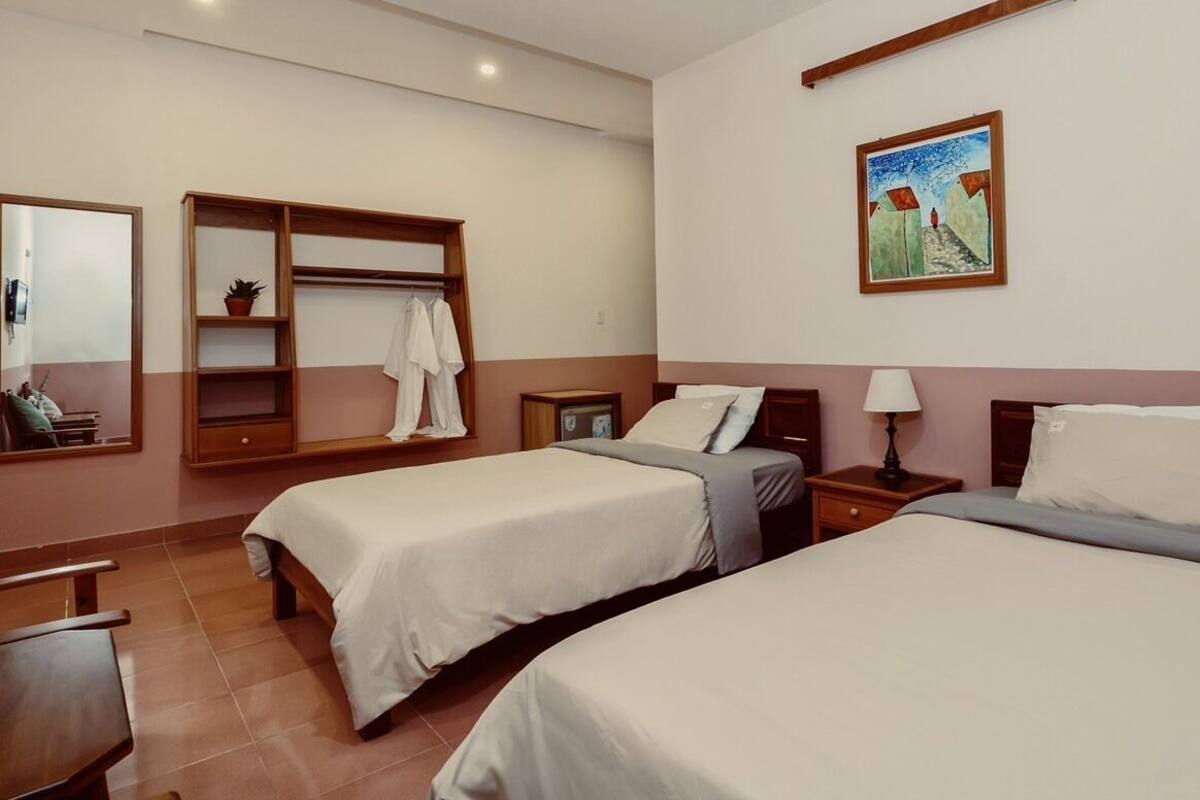 A hotel room with two beds, a wooden wardrobe, a small table, a nightstand, a lamp, a painting, and a mirror.