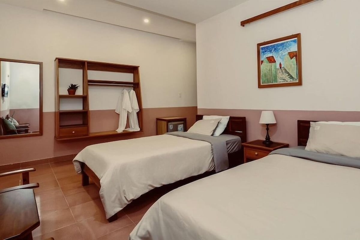 A hotel room with two beds, a wooden wardrobe, a mirror, a painting, and a small table.