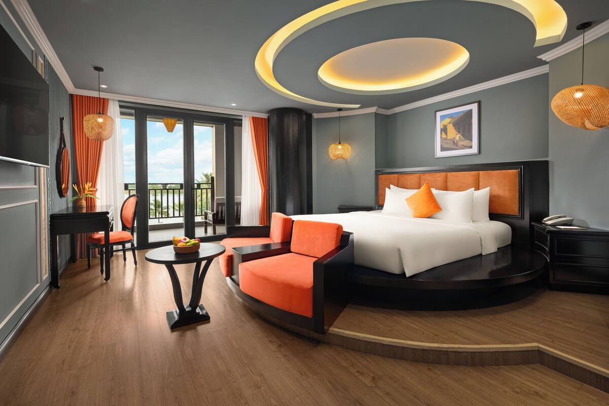 A modern hotel room with a curved bed, orange armchairs, a small table, and a balcony with a view.