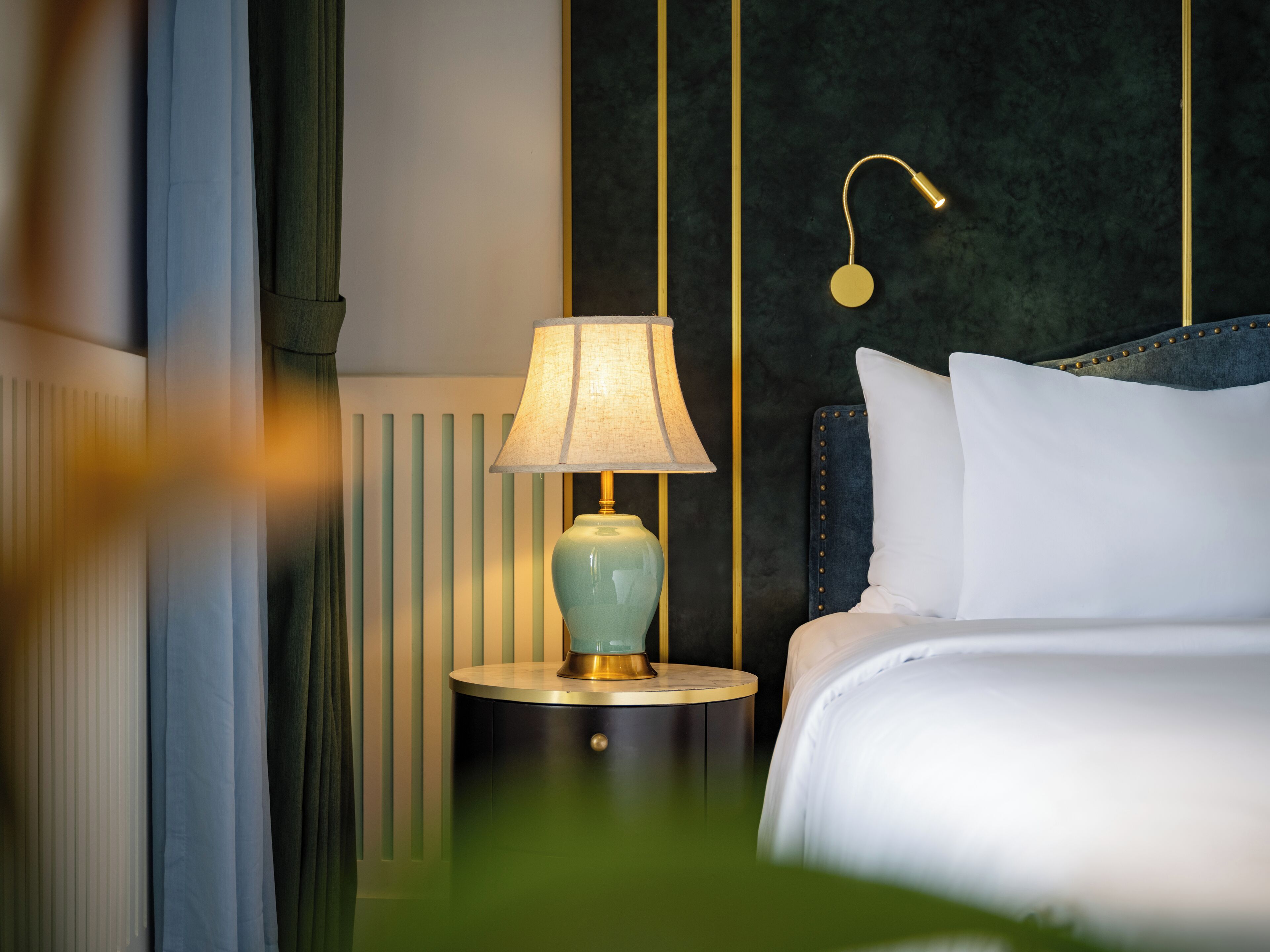 A neatly made bed with white linens, a round bedside table with a lamp, and a dark headboard with gold accents.