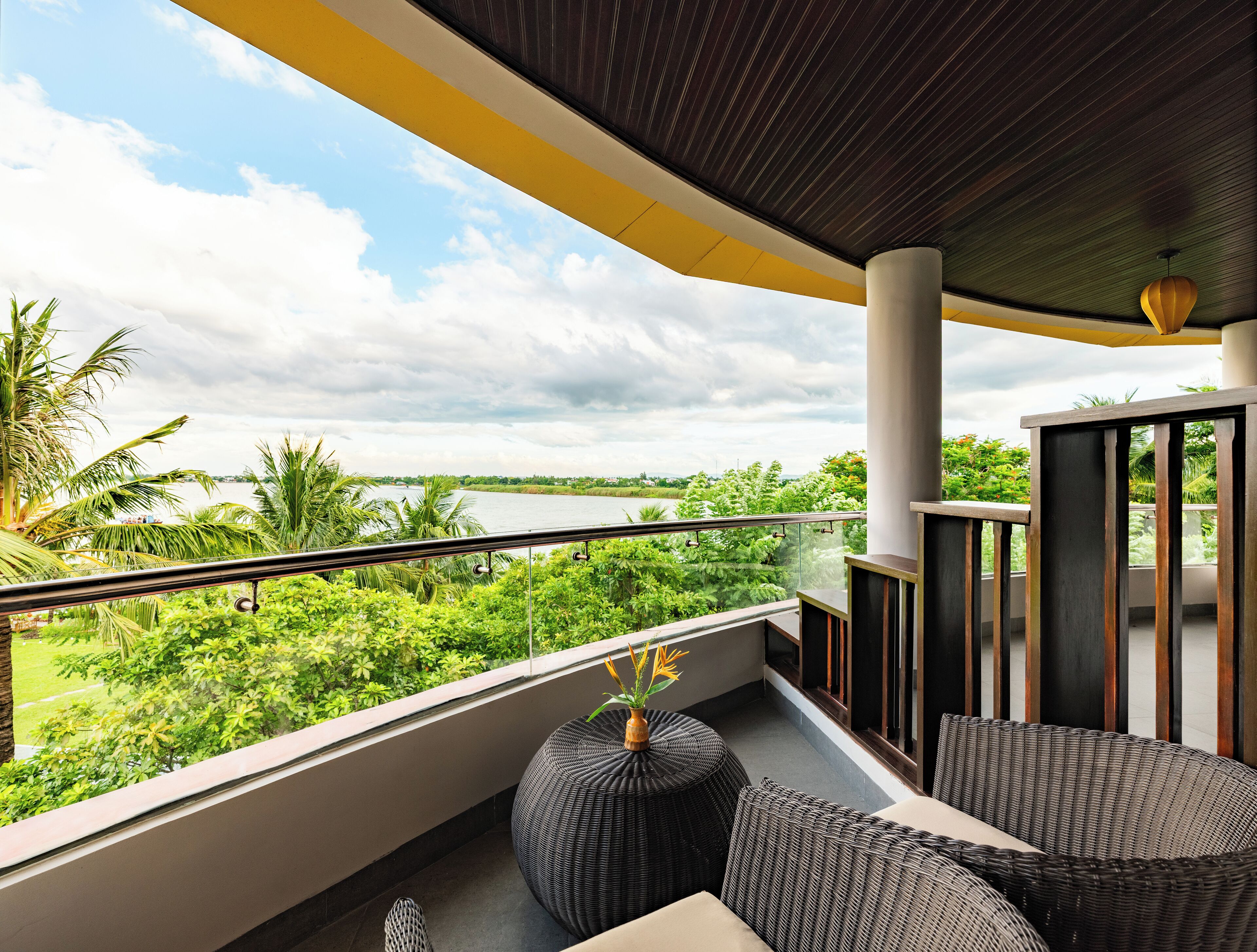 A balcony with a view of a river, surrounded by greenery, and furnished with wicker furniture.