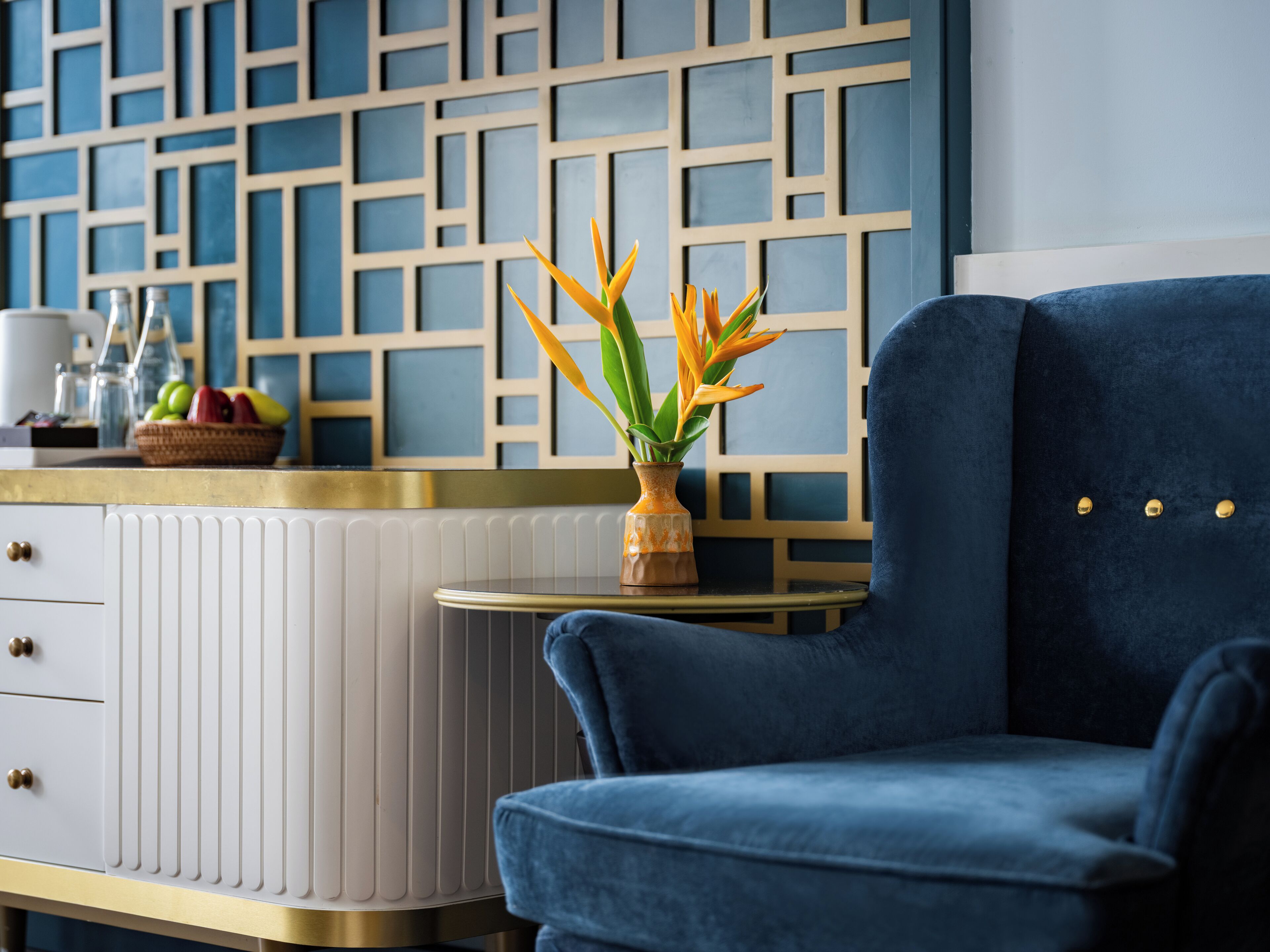 A modern living room with a blue sofa, a white cabinet with drawers, a small round table with a vase of flowers, and a decorative wall panel.