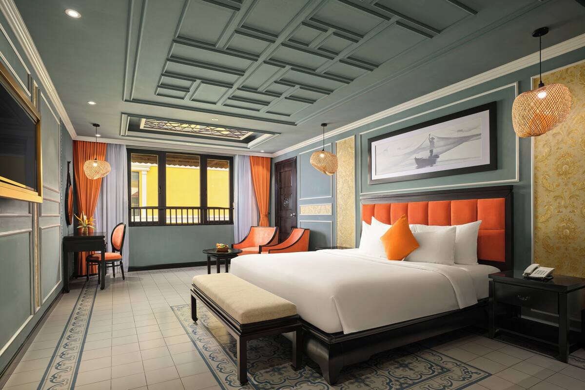 A hotel room with a large bed, a desk with a chair, a small table, and a window with curtains.