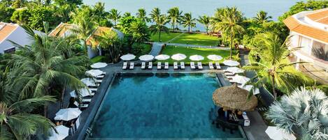 A large swimming pool with lounge chairs and umbrellas, surrounded by palm trees and a view of a body of water.