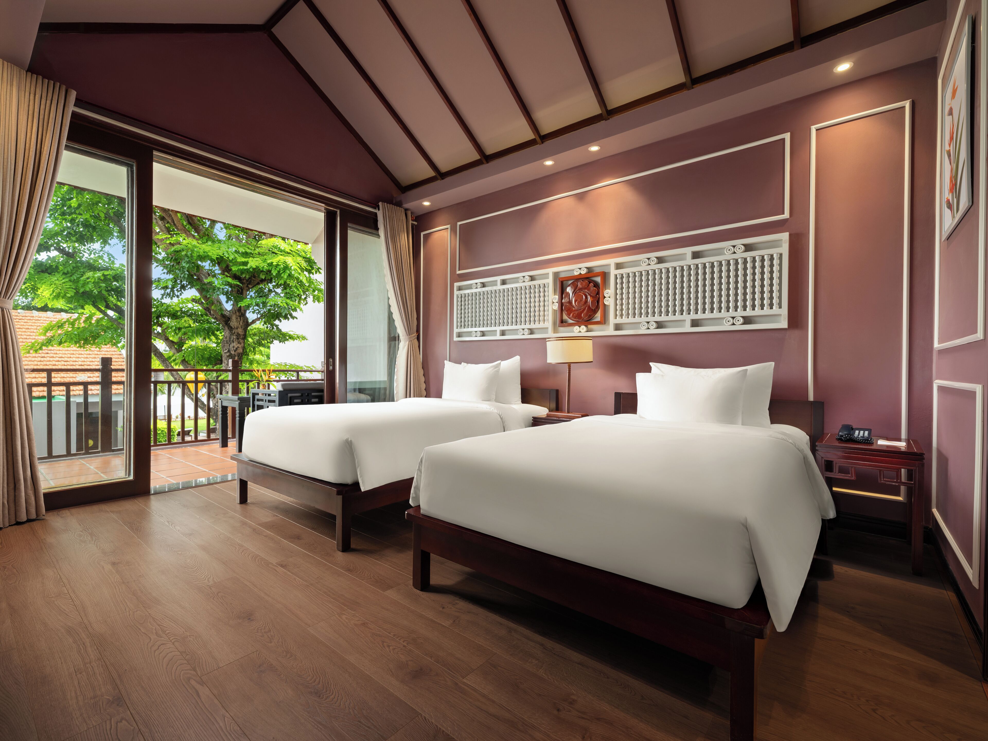 A hotel room with two beds, a large window with curtains, a wooden floor, and a view of greenery outside.