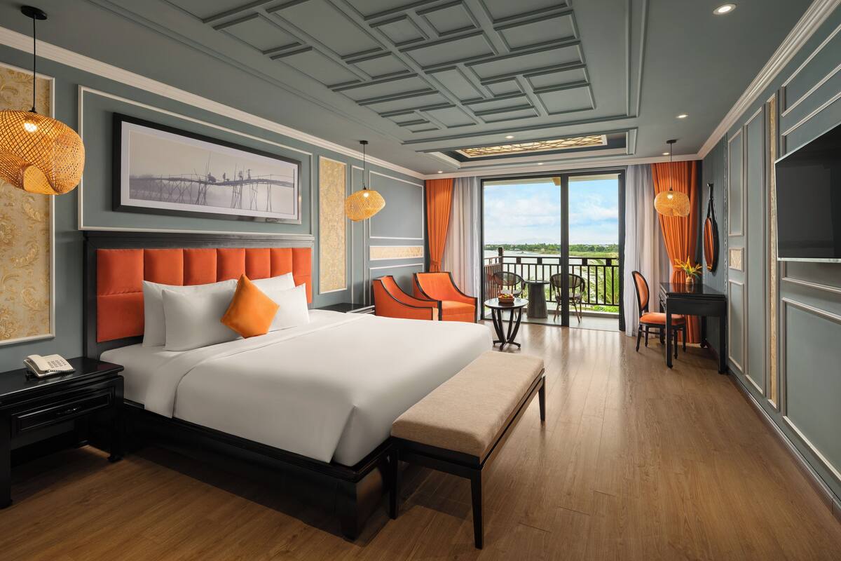 A hotel room with a large bed, a desk, two chairs, a balcony with a view, and a ceiling with a geometric pattern.