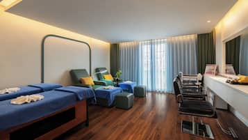 Una habitación con dos camillas de masaje, una mesa de masaje, una mesita con una planta y un ventanal con cortinas.