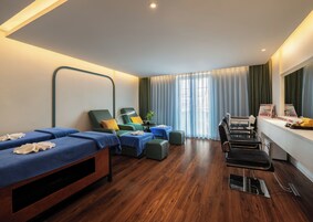 Una habitación con dos camillas de masaje, una mesa de masaje, una mesita con una planta y un ventanal con cortinas.