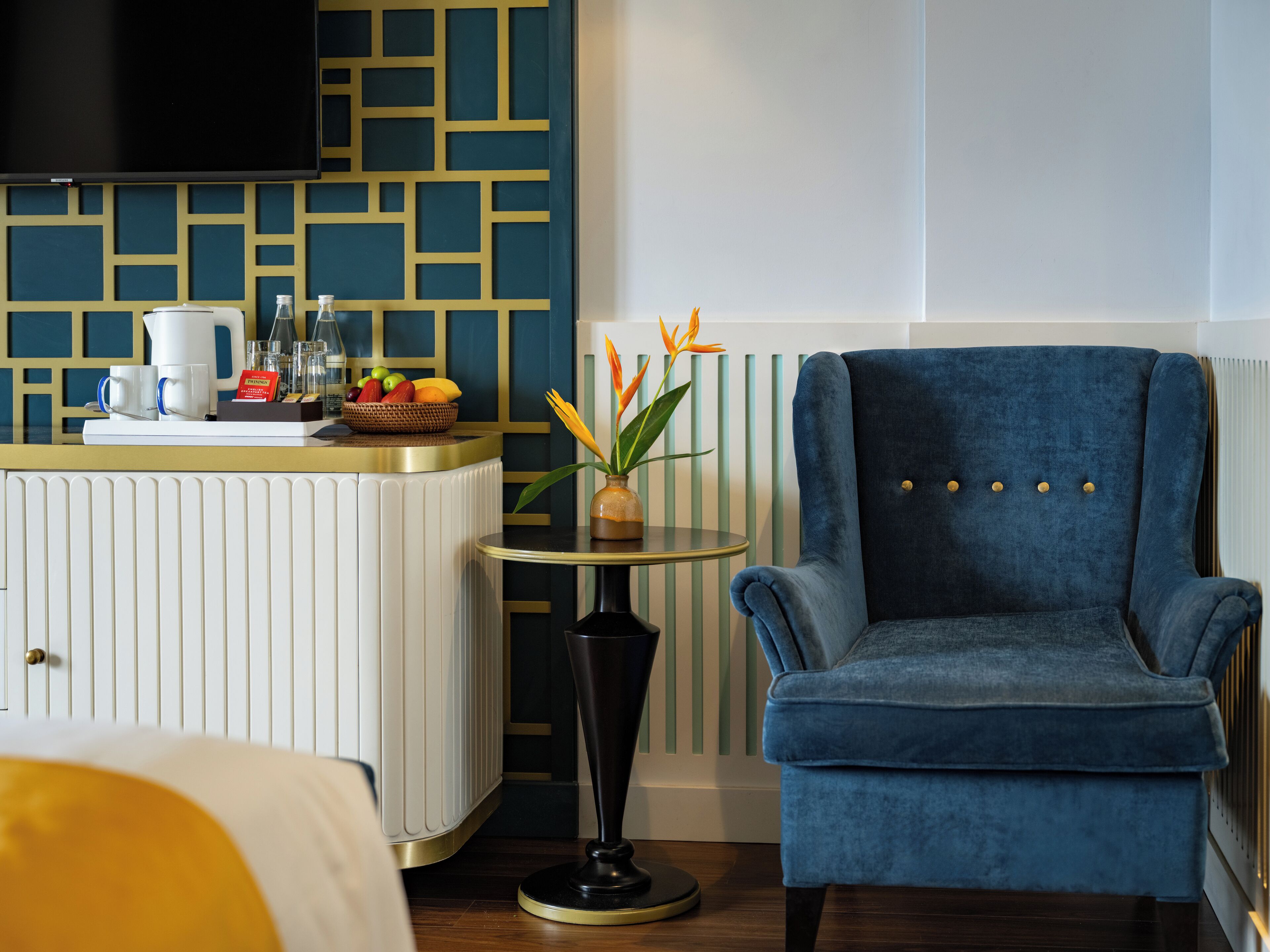 A hotel room with a blue armchair, a small round table with a vase of flowers, a fruit bowl, and a coffee maker on a counter.